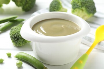 Baby food. Tasty puree in bowl, spoon, peas and broccoli on white wooden table, closeup