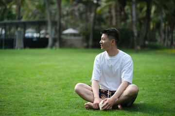 Asian man sitting cross-legged on the grass, holding a coconut and smiling sideways in Sanya, Hainan, China