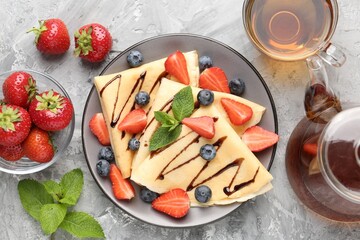 Tasty crepes with berries, chocolate sauce, mint and tea on gray textured table, flat lay