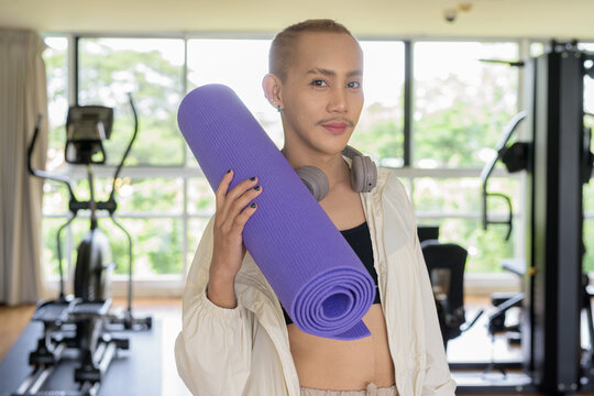 Non-binary gay man with mustache and androgynous appearance working out indoors in gym. Wearing feminine sports bra and using water bottle. Inclusive fitness concept with genderfluid representation.