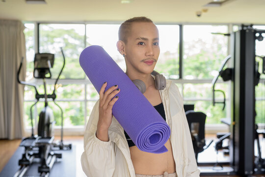 Non-binary gay man with mustache and androgynous appearance working out indoors in gym. Wearing feminine sports bra and using water bottle. Inclusive fitness concept with genderfluid representation.