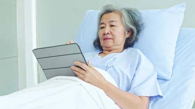 Elderly woman recovering from illness in hospital lying down to read messages on tablet digital feeling sleepy asking to take a nap on the patient bed in the afternoon.