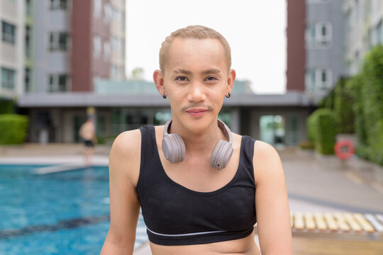 Genderfluid individual with mustache and lipstick relaxing poolside while wearing a bikini-style top. Confident, expressive LGBTQ+ representation in summer lifestyle setting.