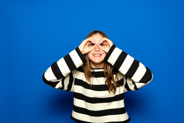 Beautiful Hispanic teenager with braces wearing a striped sweater making a thumbs-up gesture, as if holding binoculars, looking through her fingers. Fun concept