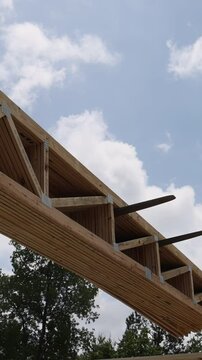Forklift telehandler machinery raises wooden trusses beams high in air as fluffy clouds fill blue sky during workday.