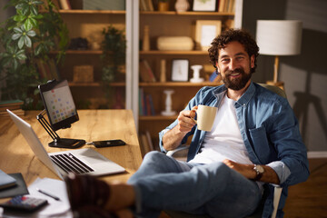 A man sits comfortably in a home office, smiling while holding a cup of coffee. His laptop and...