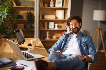 A man sits comfortably in his home office, relaxed in a chair at a wooden desk, working on a laptop while sipping coffee. The room is filled with books and greenery, creating a cozy atmosphere.