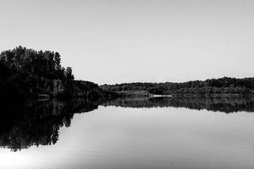 Expansive black and white landscape featuring a still lake perfectly mirroring dense forest trees...