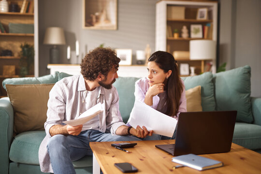 In a cozy living room, a couple engages in a serious conversation about their financial issues while examining documents and utilizing a laptop for information.