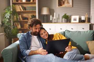 Two people sitting closely on a couch in a warm, inviting living room, sharing a joyful moment as they look at a laptop together during a relaxing evening.
