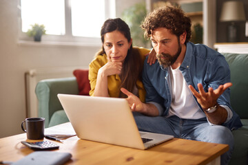 A couple appears stressed as they sit on a couch, looking at a laptop. They are surrounded by...