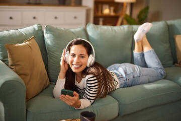 A woman lies comfortably on a green couch, enjoying music through headphones while browsing her smartphone in a warm, inviting living room setting.