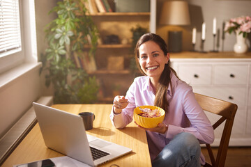 A young woman sits at a wooden table in her home office, smiling as she enjoys a bowl of cereal and...