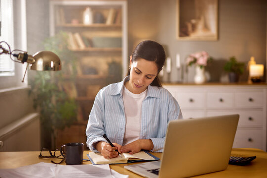 A woman sits at a wooden desk in a cozy home office, writing in a notebook with a laptop open nearby. Natural light fills the room, creating a warm and productive atmosphere. - Powered by Adobe