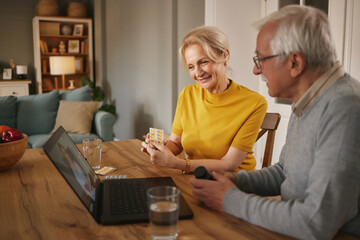 At their wooden table, a senior Caucasian couple is video chatting with their doctor. The woman is showing medicine while the man is holding a bottle. They are in their home, during the afternoon.