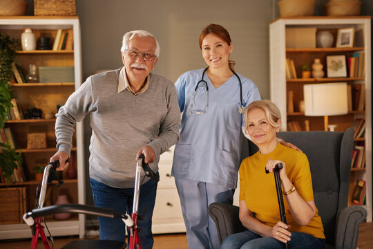 A healthcare professional in light blue scrubs and stethoscope is smiling alongside a smiling senior couple. One man is using a walker, while the woman sits in an armchair with a cane. - Powered by Adobe