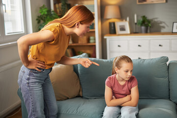 A parent is scolding a child sitting on a sofa in a cozy living room. The child looks upset,...