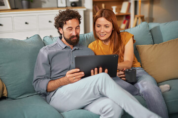 A couple sits closely on a sofa in a warm living room, happily examining a digital device. Soft...