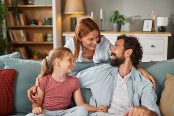 A mother, father, and daughter engage in joyful conversation on a comfortable couch in their inviting living room, surrounded by cozy decor and natural light.