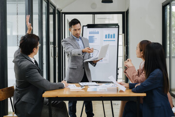 Business Presentation: A confident professional leads a presentation, holding a laptop while addressing colleagues at a boardroom table. The dynamic scene highlights collaboration, strategic thinking.