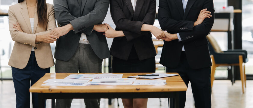 United Business Front: A group of professionals stand united at a wooden table. With their arms interlocked, they symbolize collaboration, solidarity, and partnership within a business setting. 