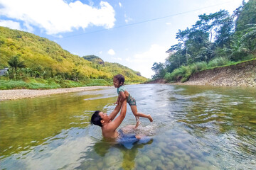 Happy Asian father and daughter enjoy playful in river with hills in background. Happy family time, outdoor adventure, bonding moment, rural nature, childhood joy, and summer activity