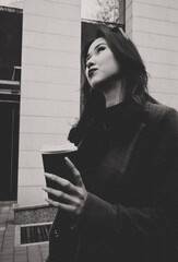 A woman in a dark coat and sunglasses holds a coffee cup, gazing upward outside a modern building...