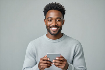 A smiling young Black man, dressed in a light grey sweater, gazes directly at the camera while holding a smartphone in his hands.