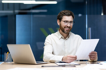 Smiling young male accountant and auditor working in the office, sitting at the table and checking...