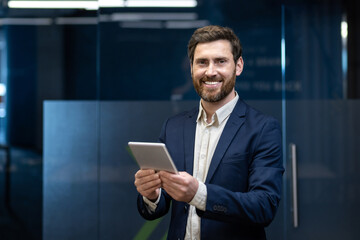Portrait of a smiling and successful young businessman in a suit standing in an office, holding a...