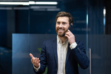 Smiling young man in a suit is standing in a company building and talking on the phone, gesturing...