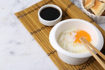Traditional asian breakfast with half-boiled egg in bowl, soy sauce, chopsticks and toasted bread on white marble table, closeup. Space for text