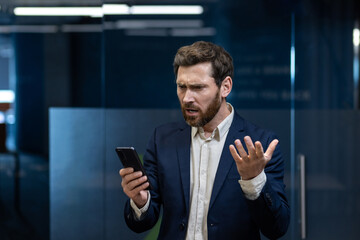 Worried young businessman in a suit standing in the office and looking at the phone screen in...