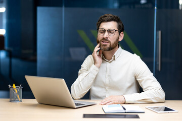 A young businessman suffering from pain sits in the office at a desk and holds his cheek with his...