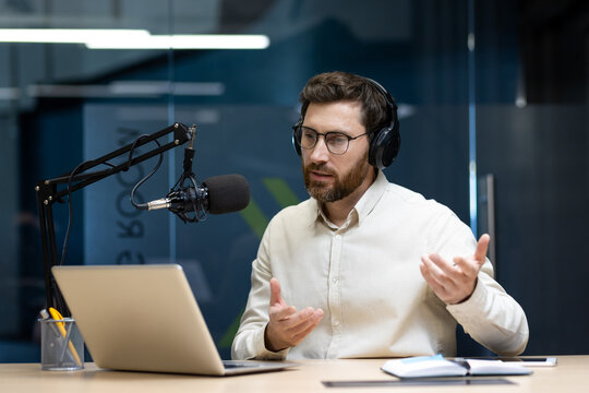 A young man wearing headphones sits in an office at a table in front of a microphone and talks via video call via a laptop, gesturing with his hands