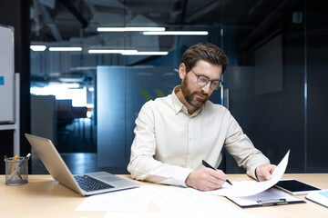 A serious and focused young male businessman is sitting in the office at his desk and working with...