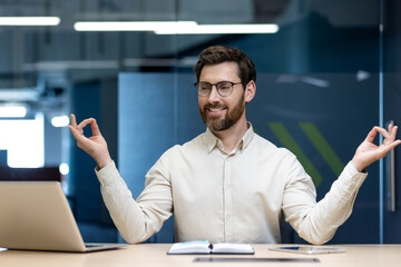 A relaxed young man works in the office, sits at his desk in the lotus position during a break and...