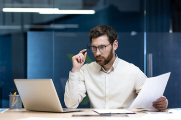 Concentrated and serious young businessman working in the office, sitting at the table, holding...