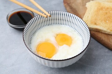 Traditional asian breakfast with half-boiled eggs in bowl, soy sauce, bread and chopsticks on grey table, above view
