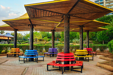 Bright circular benches painted in rainbow colors beneath umbrella-shaped wooden canopies in a...