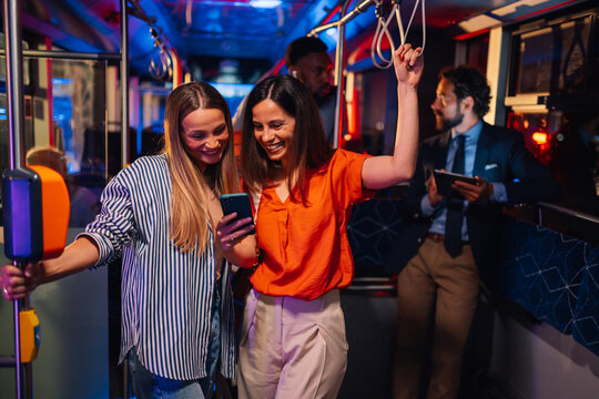 Two smiling women using smartphone on night bus ride