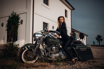 A stylish woman in a black jumpsuit poses on a vintage military motorcycle in front of a white...