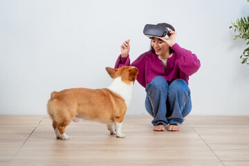 A young Asian woman relaxes indoors in an empty room, wearing VR glasses while her playful Corgi dog stays beside her. The moment captured companionship, technology, and the joy of togetherness.