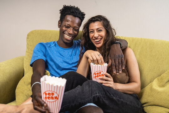 A joyful diverse couple laughing together while eating popcorn and watching a comedy movie at home.