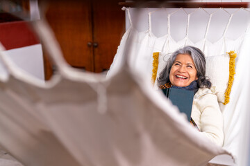 Happy senior woman relaxing in hammock, enjoying a book and smiling