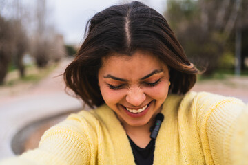 Young female influencer wearing a yellow sweater is happily laughing while recording a video blog in a park