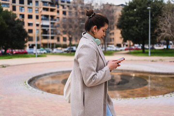 Young stylish influencer walking in a city park using her smartphone