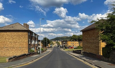 "A quiet suburban street lined with brick homes slopes gently toward the town centre, beneath a vivid sky of drifting clouds, flanked by trees and distant hills on Manor Lane, Shipley, Yorkshire, UK."