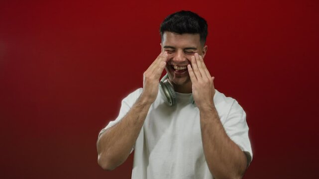 Young man with headphones covering face and laughing joyfully against vibrant red background, expressing happiness and emotion.