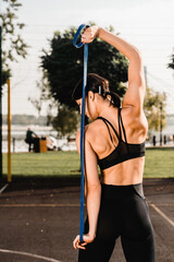 Rare view of fitness girl. Workout with resistance band outdoor. Fit woman with strong muscular body in sporty outfit doing exercise against sports park court. Active lifestyle for urban people.